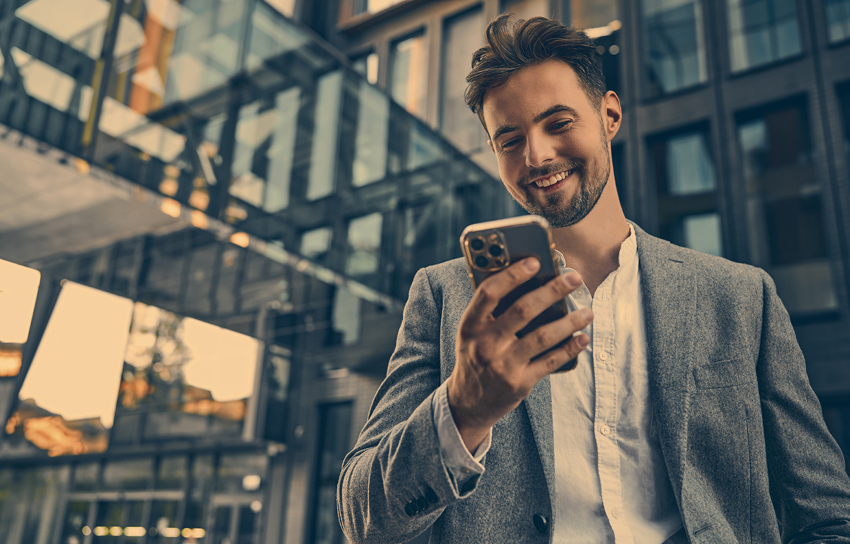 Portrait of attractive confident modern smiling happy adult dressed gray suit man holding a phone and standing outside near office building. Online work, communication. Copy space.